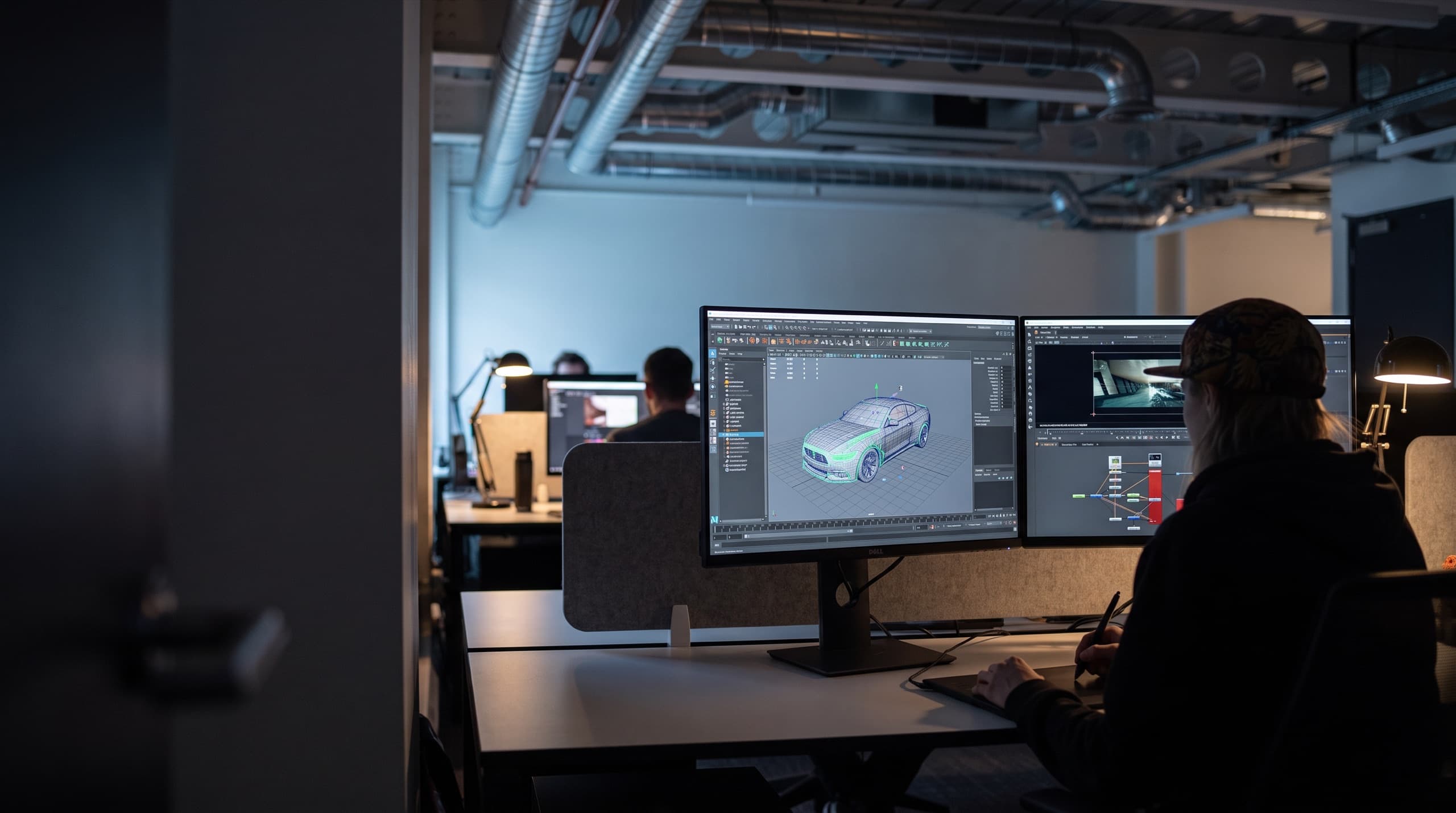 A CG artist at two monitors in a darkened studio, a car model on one screen and a compositing node graph on the other.